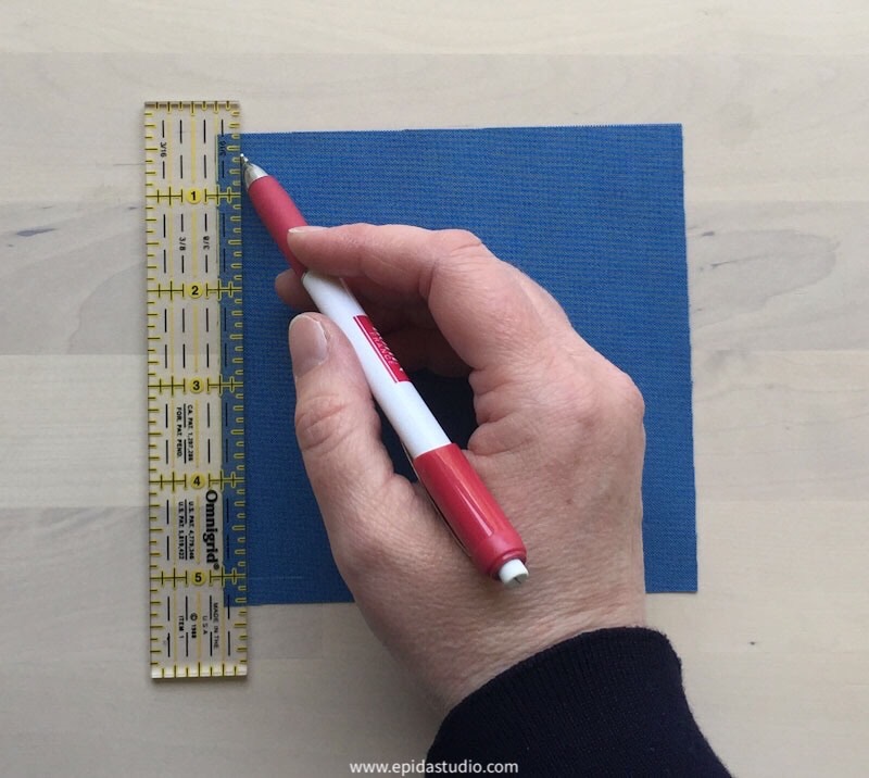 hand marking a 1/4" seam allowance on a square of blue fabric.